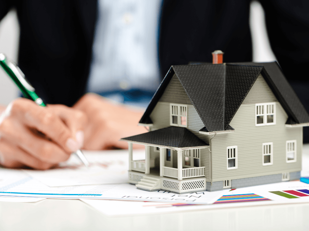 Miniatura de residência sobre documentos enquanto um profissional escreve, ilustrando a avaliação e negociação da comissão imobiliária.