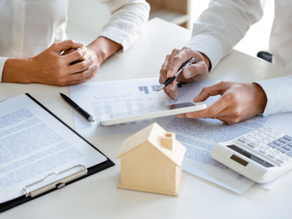 Pessoa analisa documentos, tablet e calculadora sobre uma mesa com miniatura de casa, representando o cálculo da comissão imobiliária. Tudo para explicar qual a porcentagem da imobiliária na venda de um imóvel.