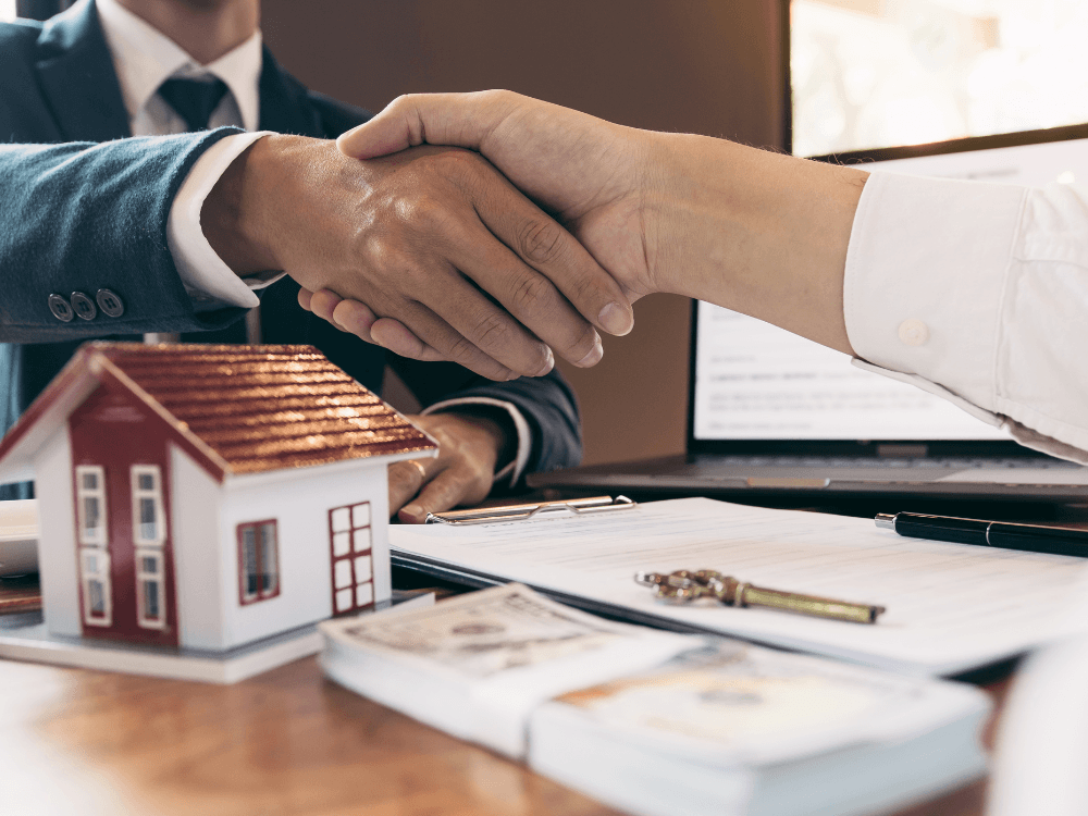 Duas pessoas apertam as mãos sobre uma mesa com documentos, dinheiro e miniatura de casa após acordo de venda imobiliária.