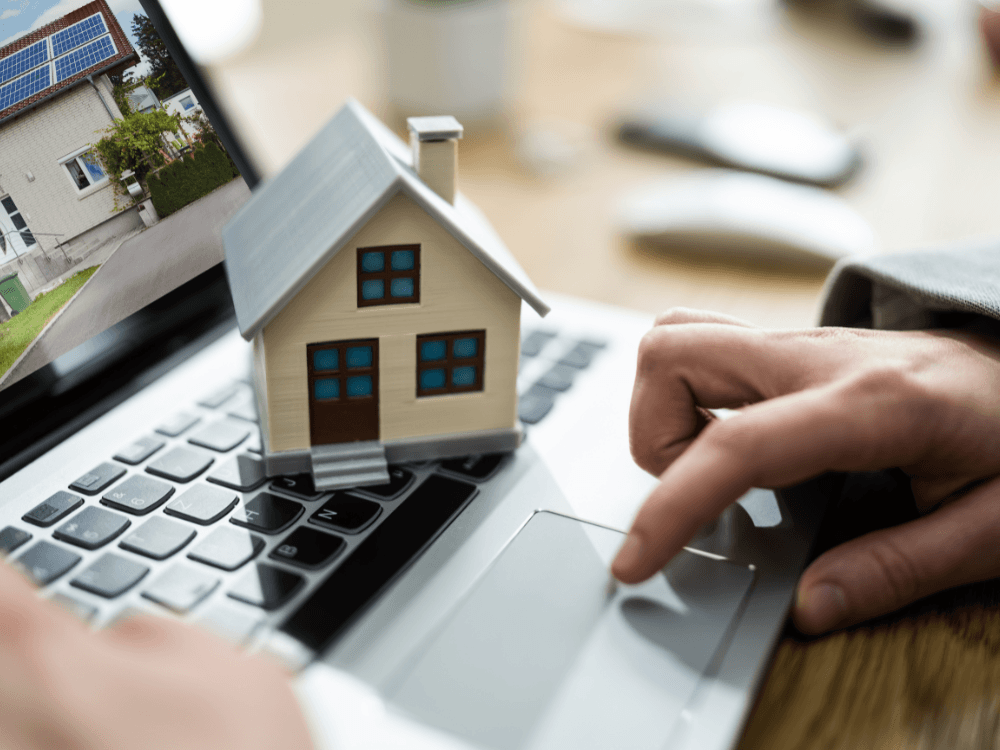 Miniatura de uma casa sobre um notebook, com imóveis ao fundo e uma pessoa pesquisando opções para compra assistida de imóvel. Tudo para explicar o que é compra assistida de imóvel.