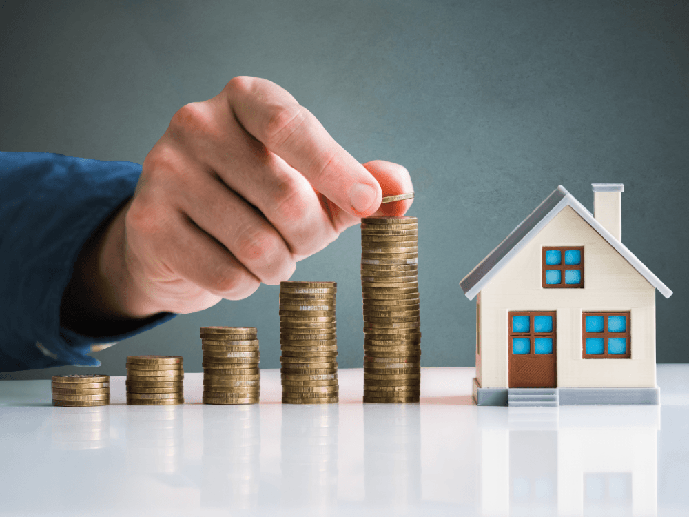 Maquete de casa ao lado de pilhas crescentes de moedas e uma mão adicionando dinheiro, ilustrando valorização imobiliária.
