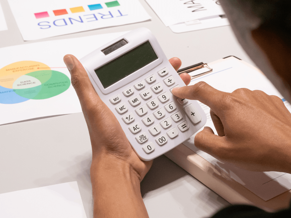 Pessoa utiliza uma calculadora sobre documentos e gráficos financeiros para realizar cálculos relacionados a despesas e valores contratuais.